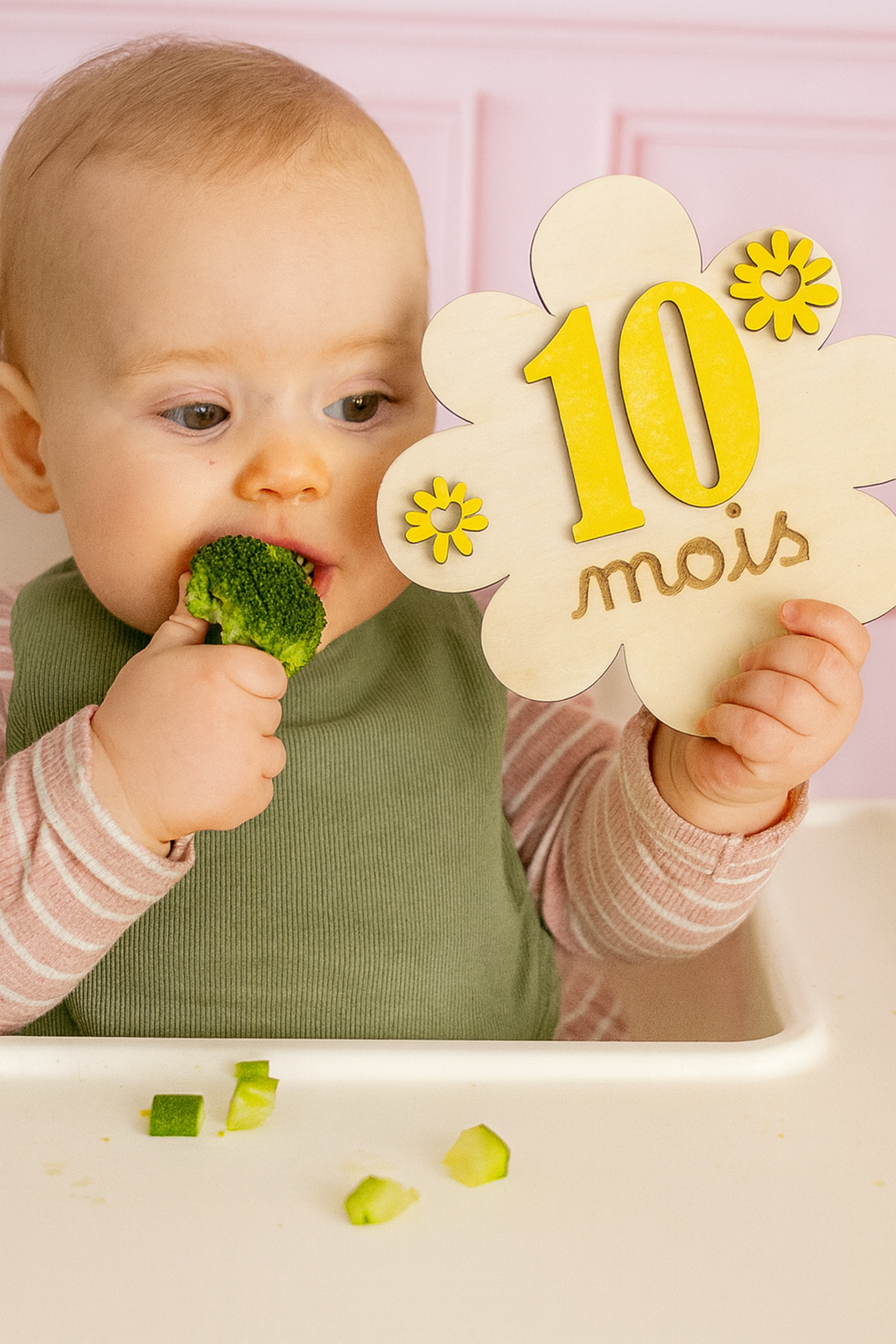 bébé assis mange un brocolis et tient une carte en bois 10 mois en forme de fleur