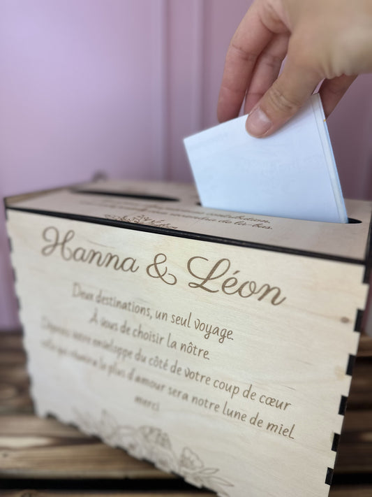 main glissant un chèque dans urne mariage en bois personnalisée "Hanna & Léon" et message gravé personnalisé.IL y a 2 entrées pour voter pour destination lune de miel. le fond est rose et la table sur laquelle est posée l'urne est en bois massif