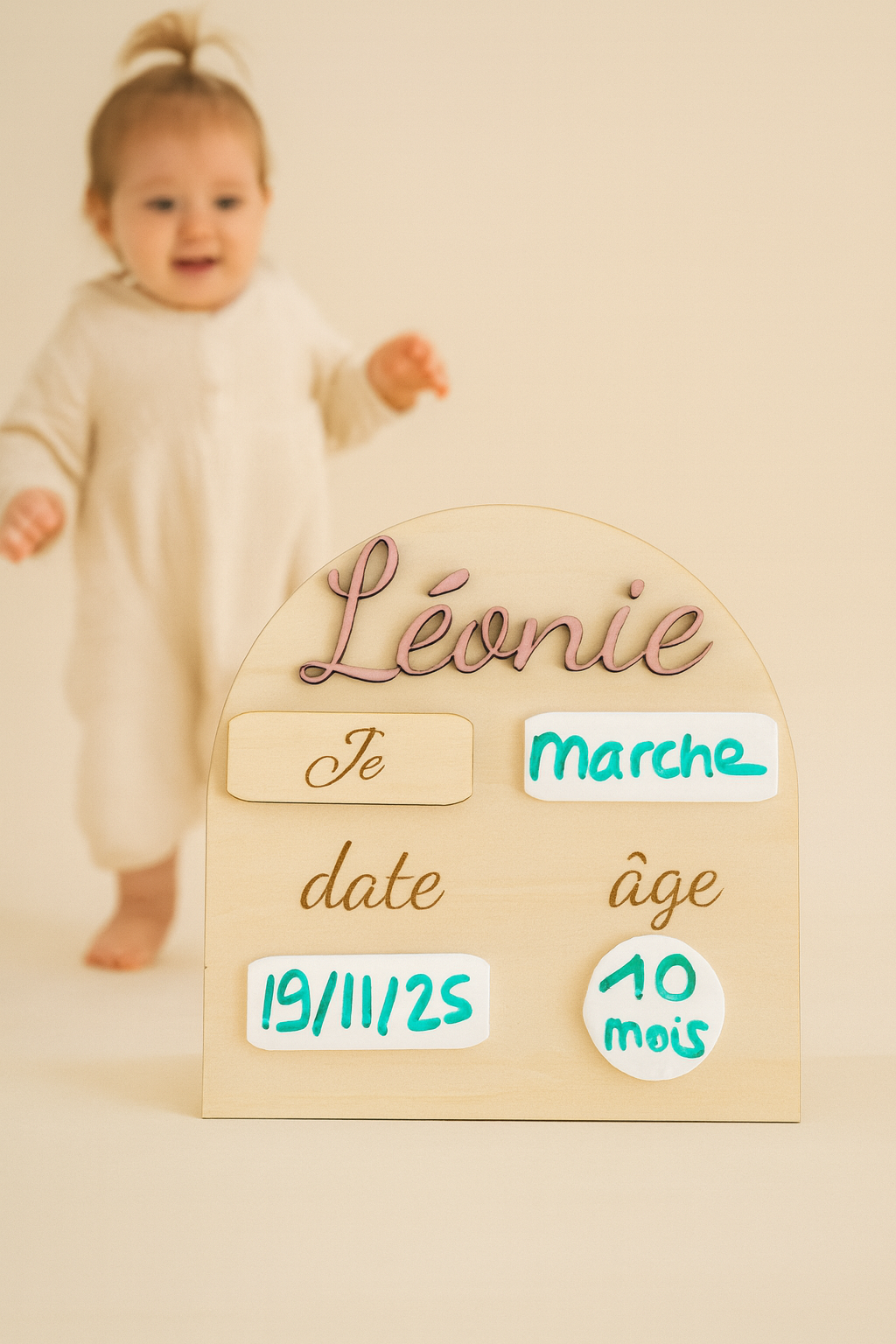Pancarte en bois personnalisée pour bébé, portant le prénom « Léonie » peint en rose, avec des zones en plexi blanc effaçable pour noter la date et l’âge. Un bébé marche vers la plaque « Je », et sur le plexi écrit "marche" ainsi que la date et l'âge. sur un fond clair et lumineux.