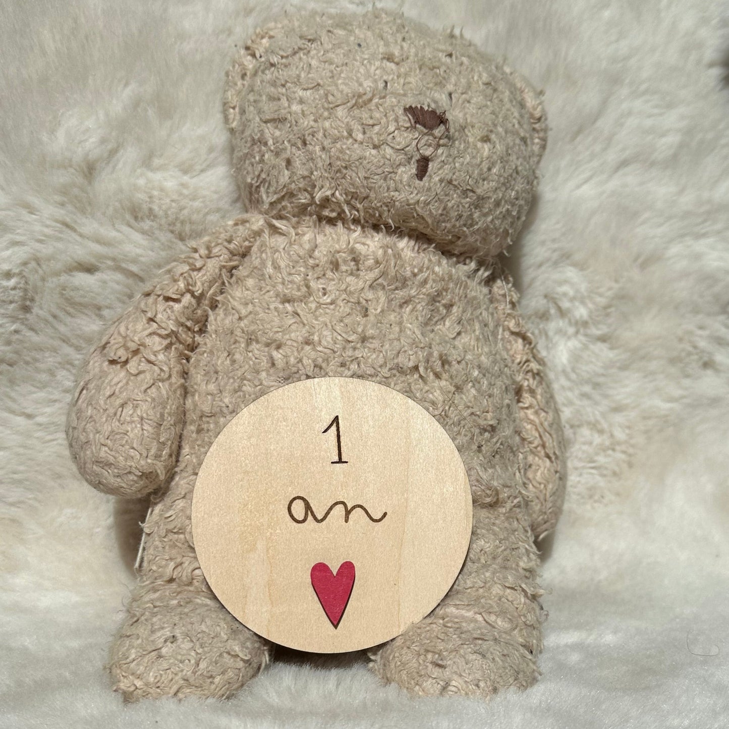 carte étape ronde en bois "1 an" avec petit coeur rose collé dessus. Carte posée sur l'ours en peluche Moonie