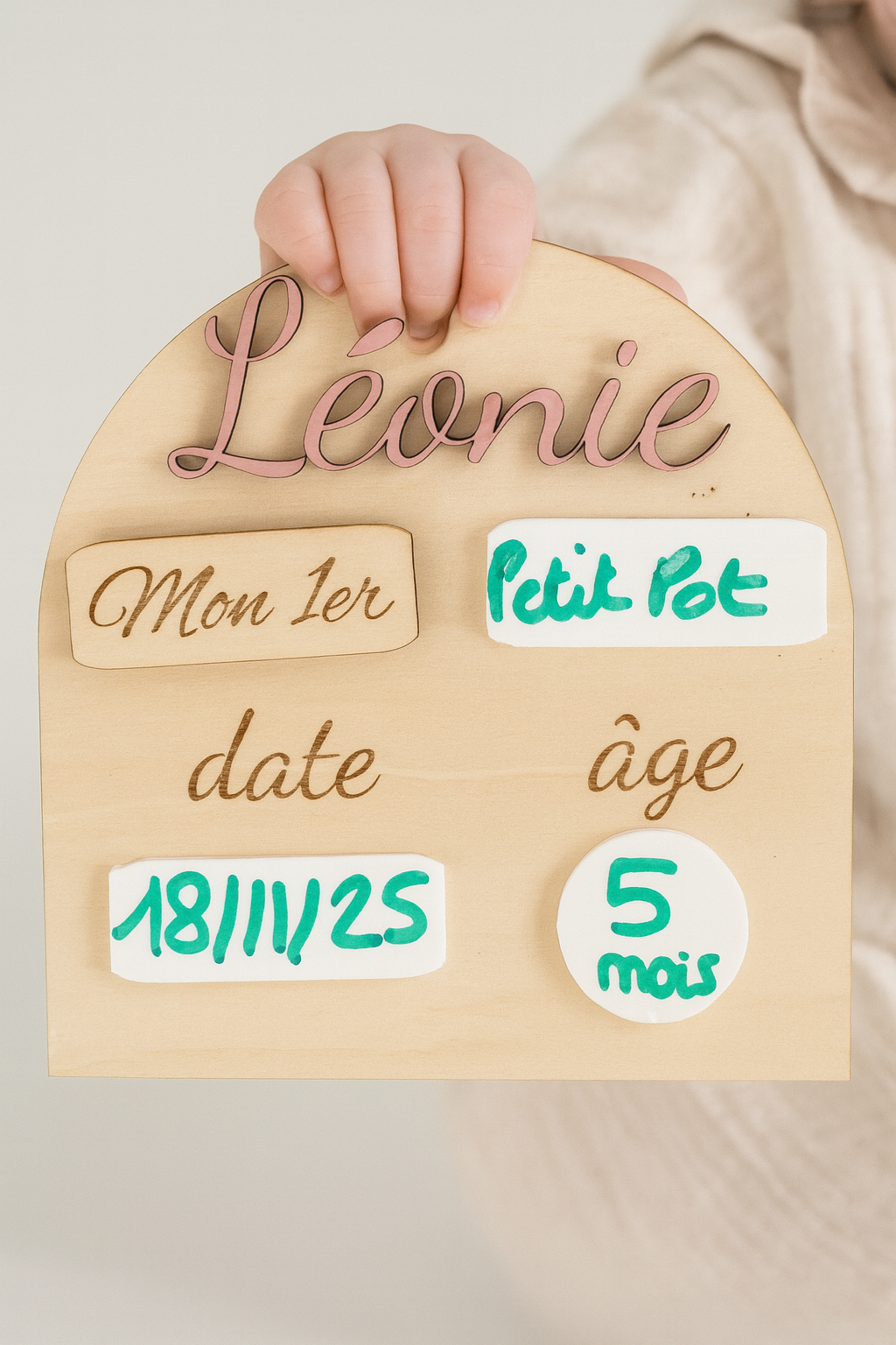 Pancarte en bois personnalisée pour bébé, portant le prénom « Léonie » peint en rose, avec des zones en plexi blanc effaçable pour noter la date et l’âge. Une main tient la petite plaque gravée « Je », interchangeable avec « Mon 1er », sur un fond en bois clair et lumineux.