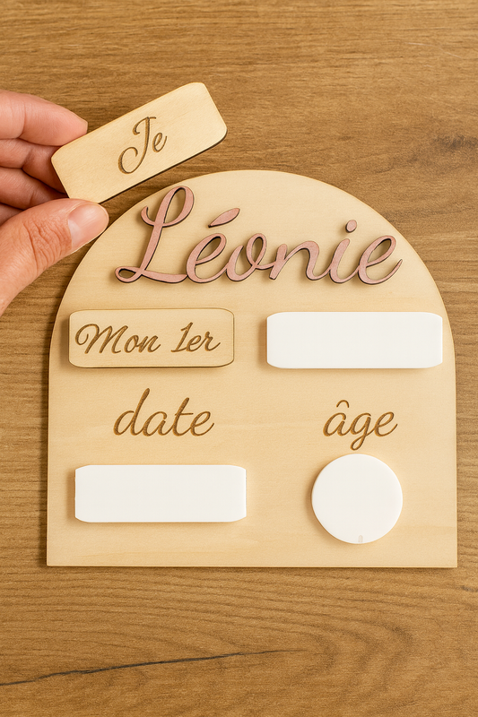 Pancarte en bois personnalisée pour bébé, portant le prénom « Léonie » peint en rose, avec des zones en plexi blanc effaçable pour noter la date et l’âge.  « Je », interchangeable avec « Mon 1er », sur un fond en bois clair et lumineux.