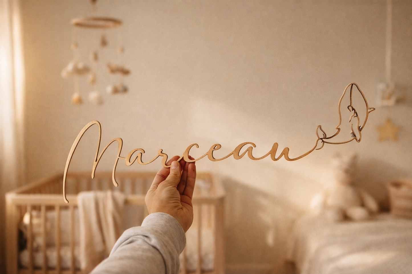 prénom en bois "Marceau" se terminant par une baleine sur fond d'une chambre d'enfant tenue par une main