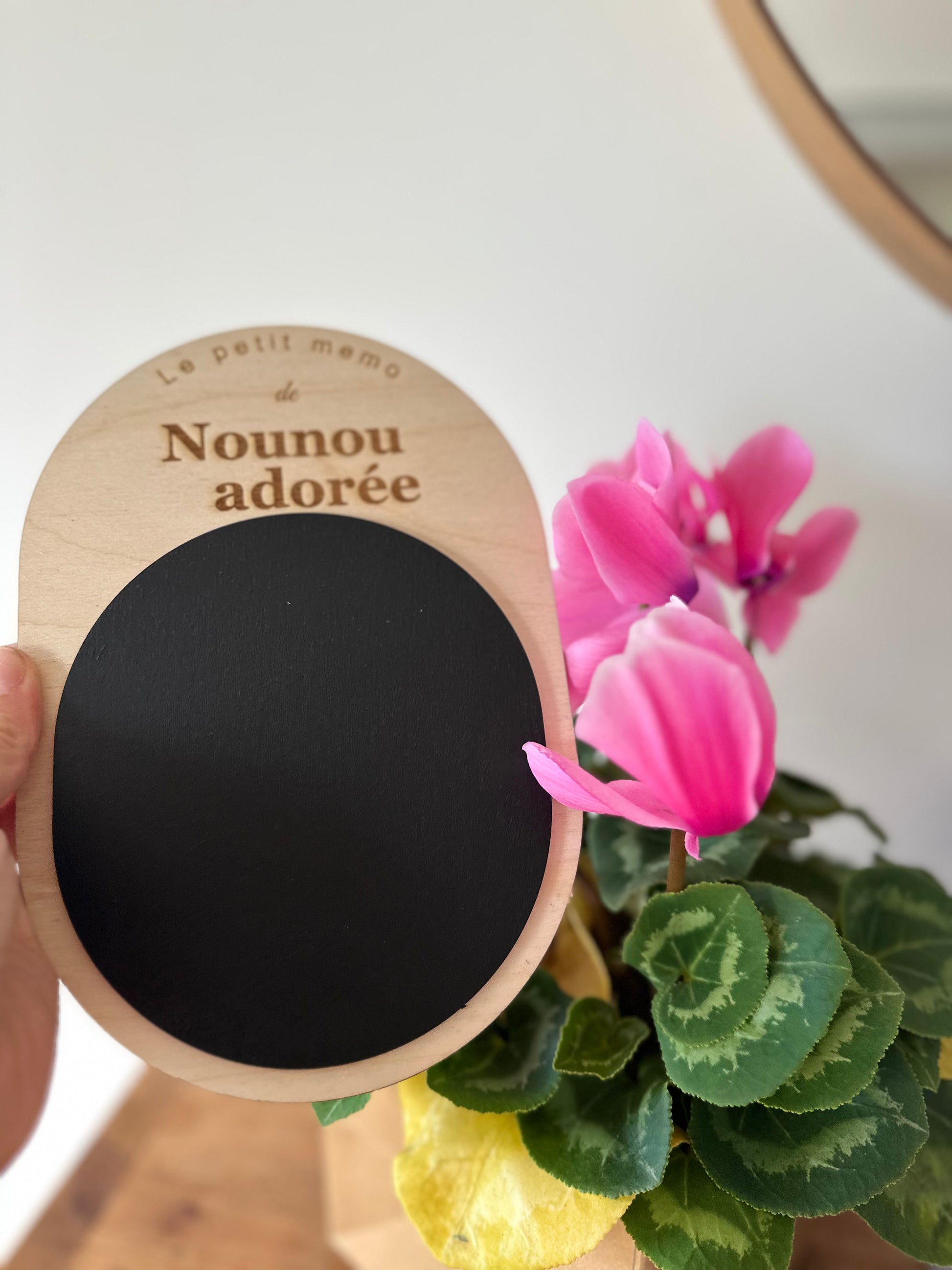 Cadeau nounou de fin d'année personnalisé : mémo en bois gravé « Nounou adorée » avec ardoise noire, tenu à la main près d'un cyclamen rose