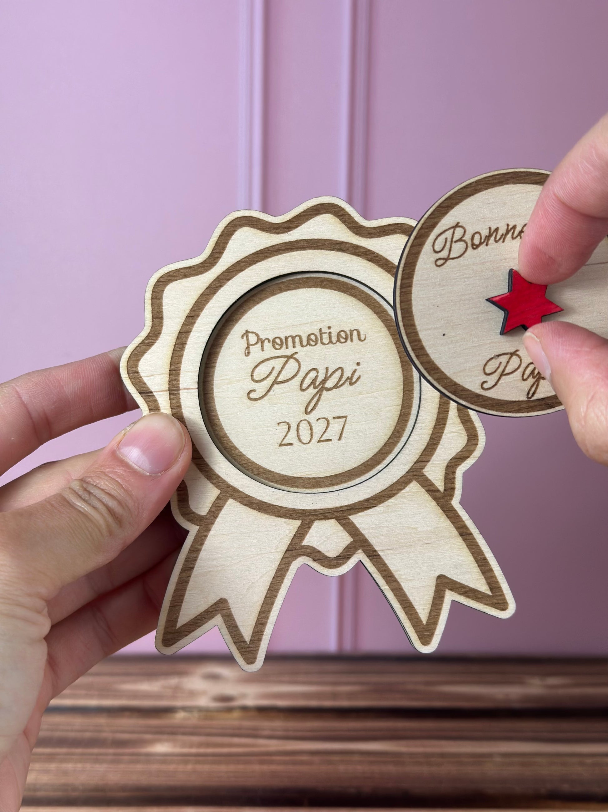 annonce grossesse Promotion papi 2027 en forme de Médaille en bois gravée 
avec couvercle Bonne fête Papa à soulever - Chou Rose, tenue par une main féminine, fond : mur rose poudré, table : boid massif