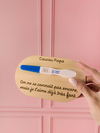 annonce grossesse coucou papa personnalisé avec un message gravé "on ne se connait pas encore, mais je t'aime déjà très fort" et test de grossesse collé, mur rose poudré, carte tenue par une main féminine