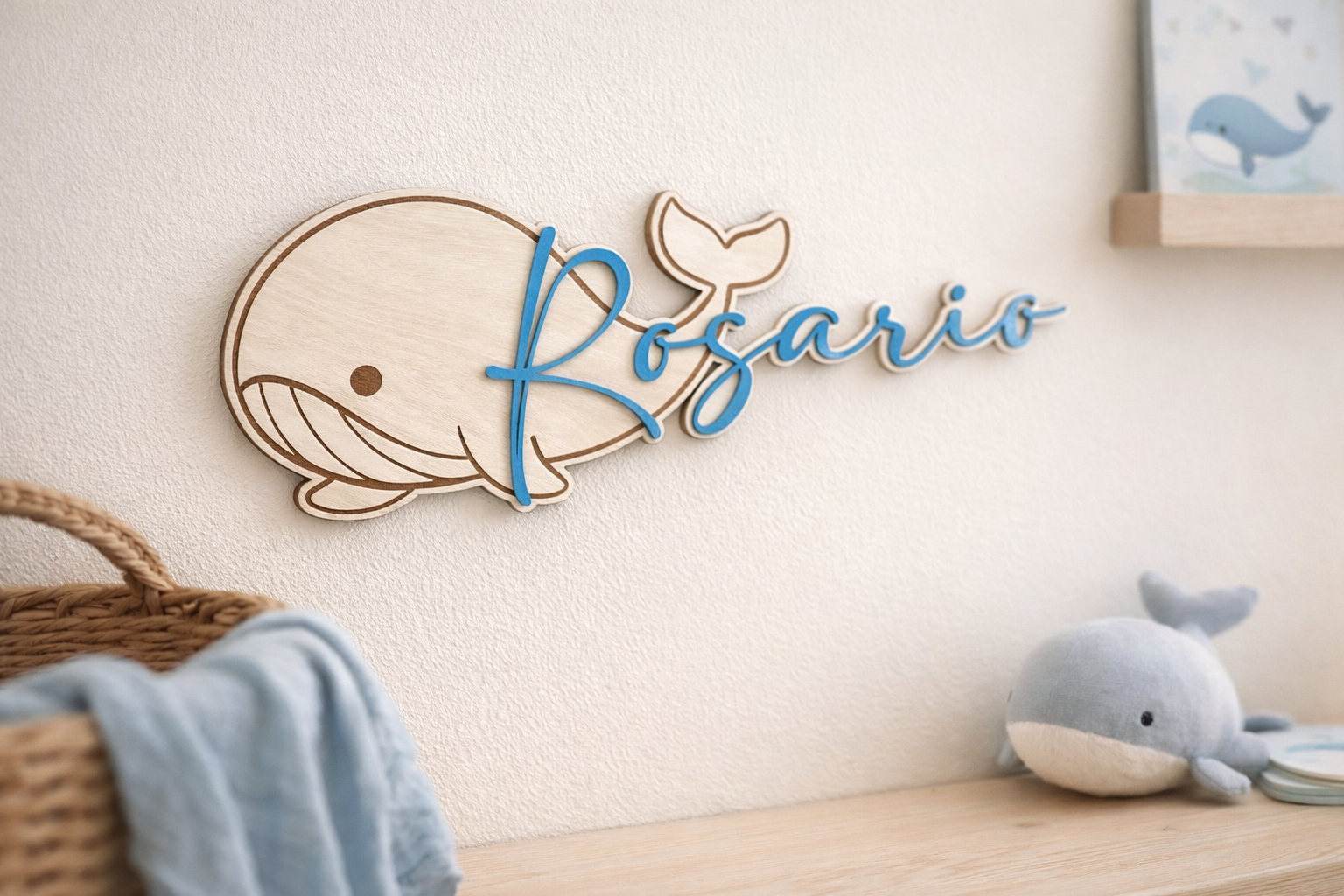 prénom en bois peint en bleu "Rosario" et fixé sur une baleine en bois gravée pour une décoration de chambre sut l thème marin. La baleine est fixée sur un mur blanc, au dessus d'une étagère sur laquelle est posée une peluche baleine, un panier dont sort un linge bleu. Sur le c^pité on aperçoit une illustration de baleine posée sur une petite étagère en bois