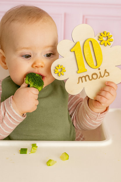 bébé assis mange un brocolis et tient une carte en bois 10 mois en forme de fleur