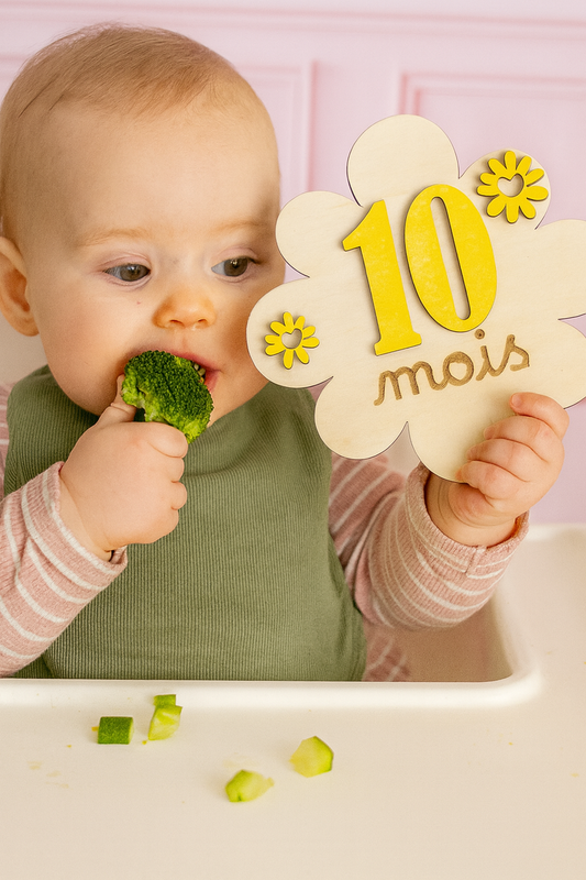 bébé assis mange un brocolis et tient une carte en bois 10 mois en forme de fleur