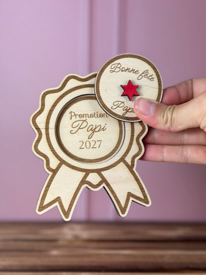 médaille en bois pour une annonce grossesse promotion Papi 2027 avec couvercle Bonne fête Papa à soulever grâce à une étoile rouge - Chou Rose, tenue par une main féminine, fond : mur rose poudré, table en bois