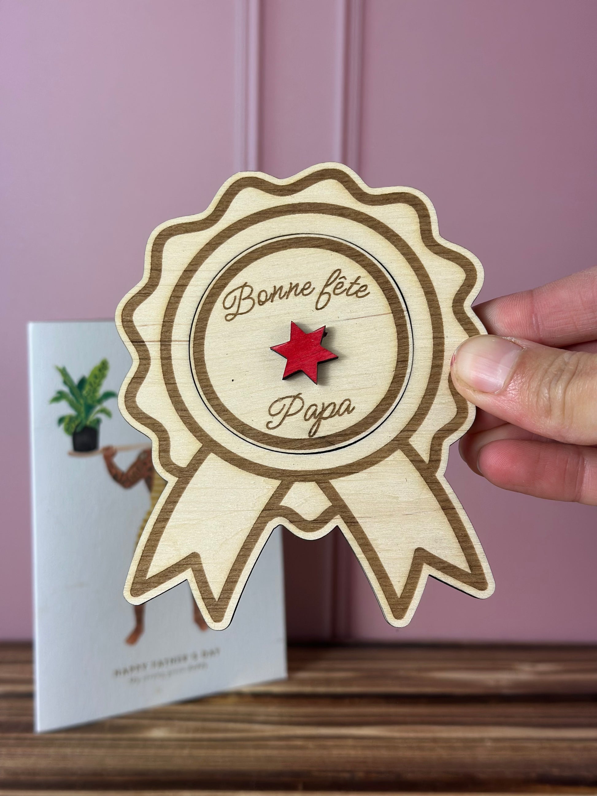 annonce grossesse en forme de méfdaille avec message gravé personnalisé à découvrir. en arrière plan, : un carte fête des pères et un mur rose