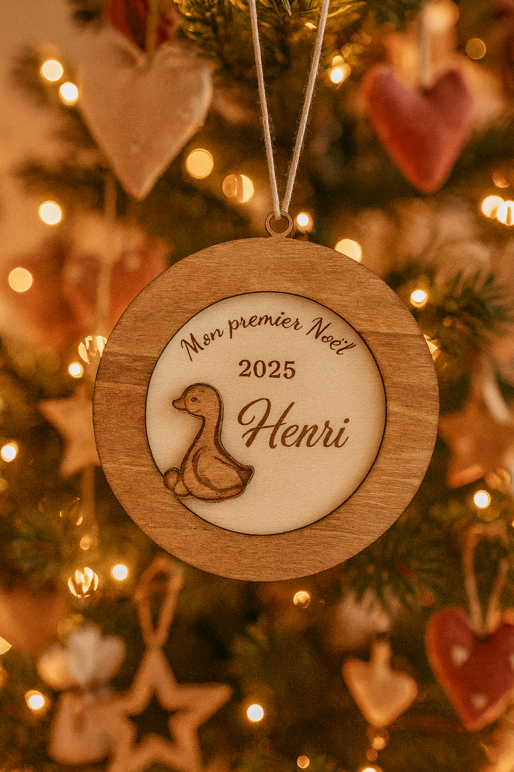 suspension ronde en bois en 3 dimensions : gravé "mon premier noël" puis "2025" et enfin le prénom de l'enfant "Henri" et détail d'un petit caneton vernis. La boule de noël personnalisée est accroché dans un sapin illuminé