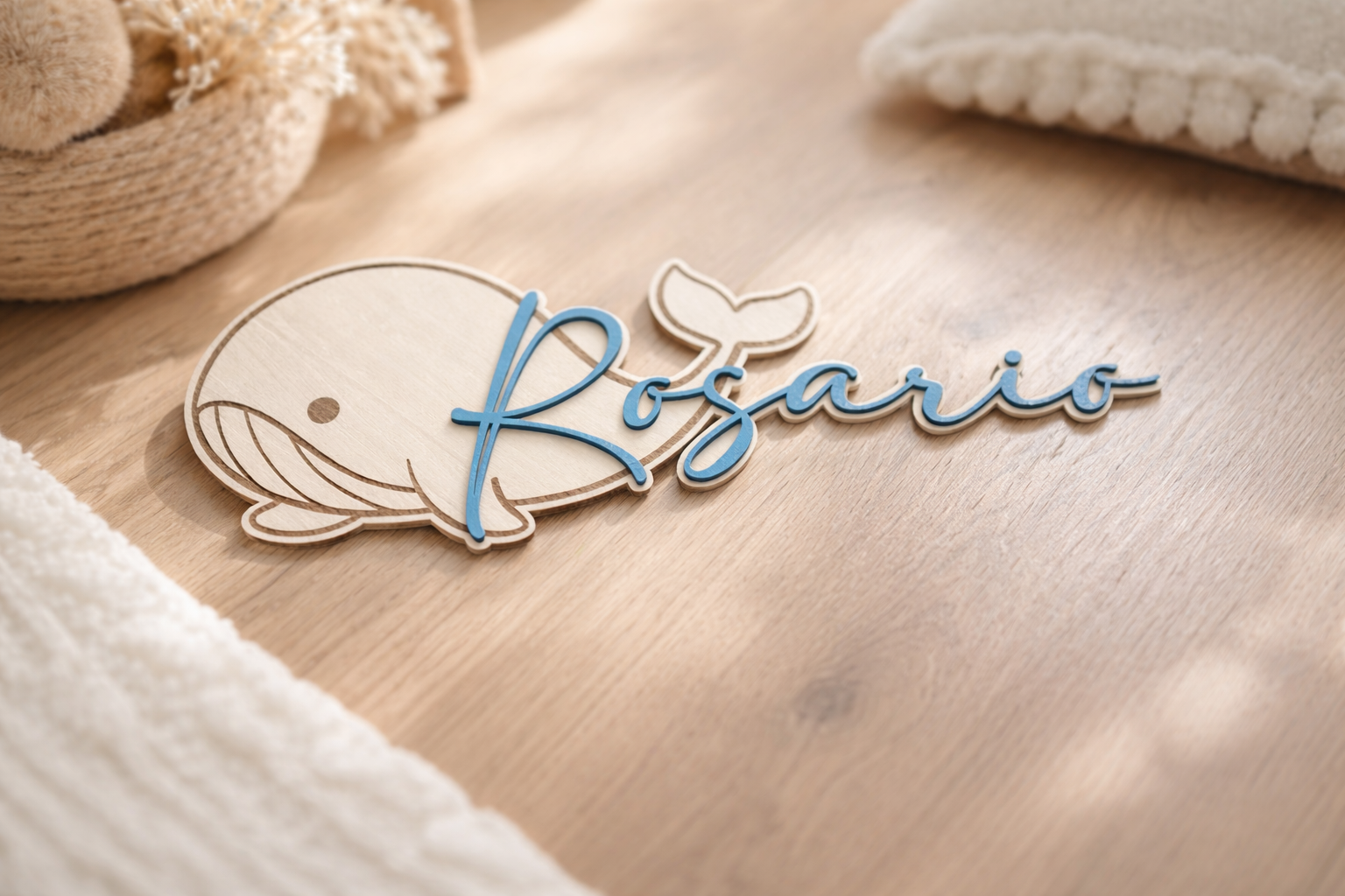 prénom en bois peint en bleu "Rosario" et fixé sur une baleine en bois gravée pour une décoration de chambre sut l thème marin. La création est posée sur une commode en bois, prêt d'un coussin à pompons écrus, d"un linge blanc et d'un panier en osier. La photographie évoque douceur, tons naturels. La scène est illuminé naturellement par les lueurs du soleil