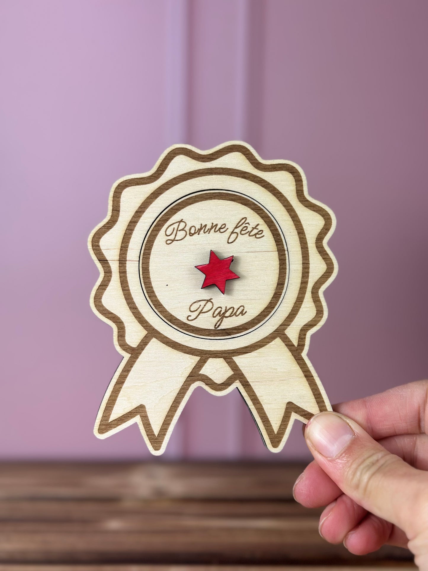 annonce grossesse Papi 2027 en forme de Médaille en bois gravée 
avec couvercle Bonne fête Papa à soulever - Chou Rose, tenue par une main féminine, fond : mur rose poudré, table : boid massif