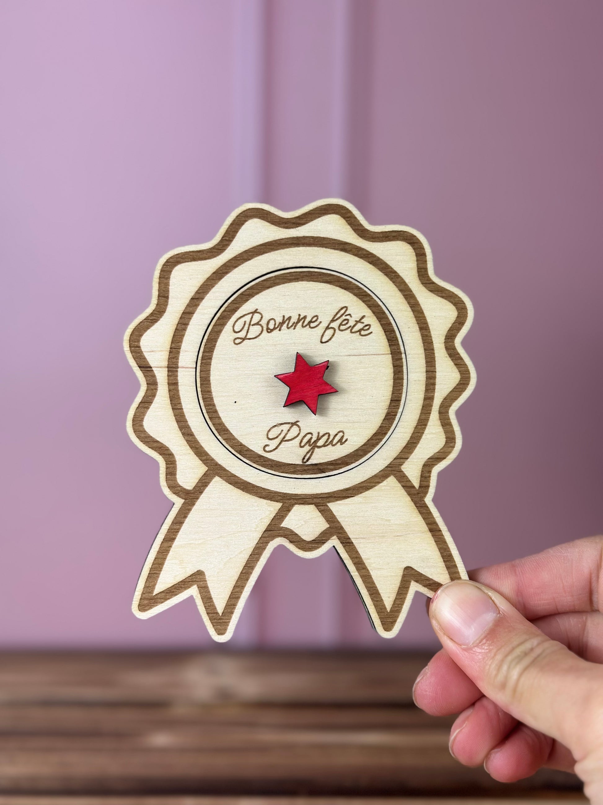 annonce grossesse Papi 2027 en forme de Médaille en bois gravée 
avec couvercle Bonne fête Papa à soulever - Chou Rose, tenue par une main féminine, fond : mur rose poudré, table : boid massif