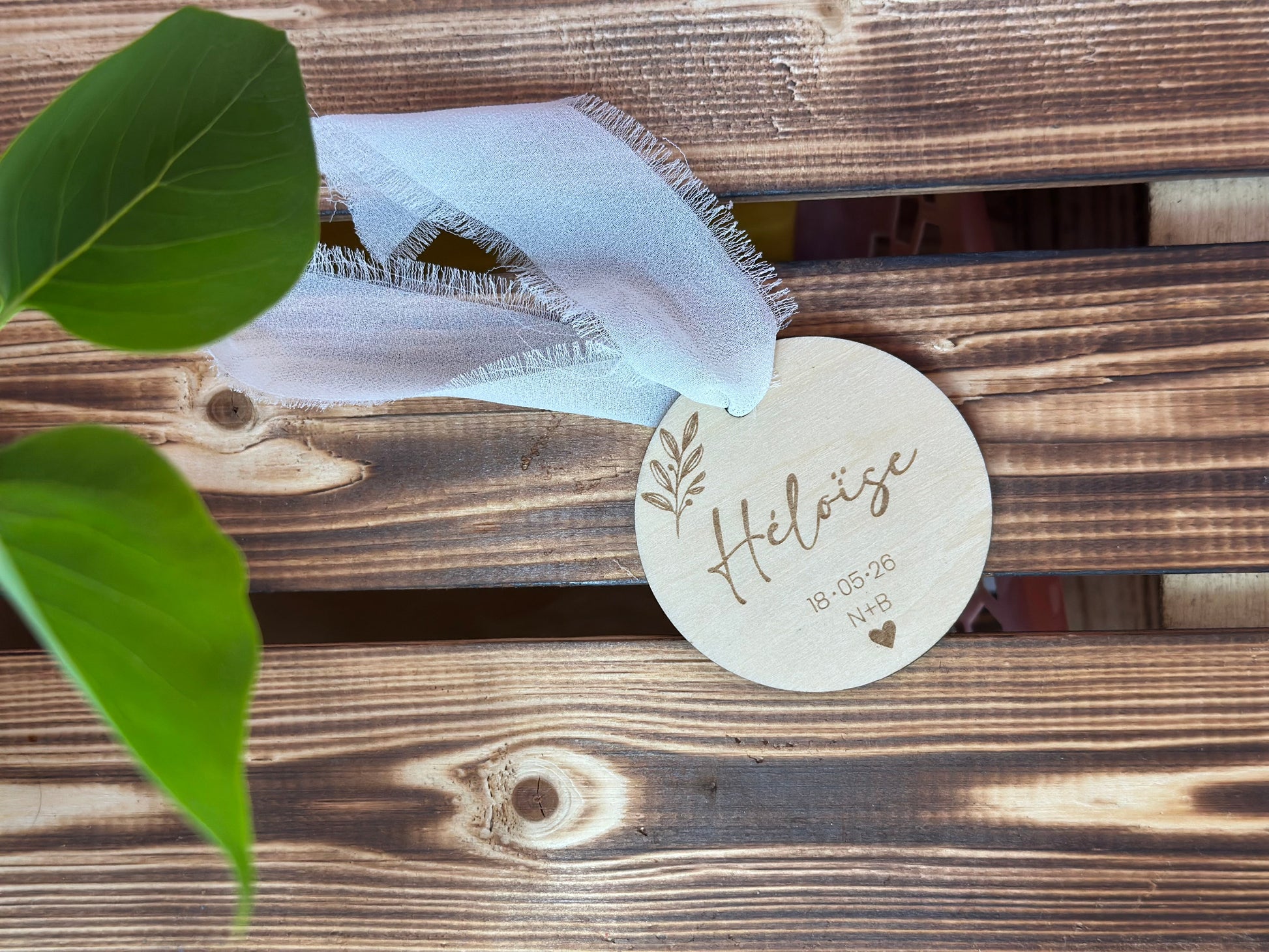 un marque place en bois personnalisé avec le prénom de l'invité gravé, la date du mariage, les initiales des mariés et des motifs végétaux. Le cadeau invité est posé sur une palette en bois et orné d'un ruban
