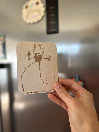 une main tient le dessin de votre enfant reproduit sur une carte en bois