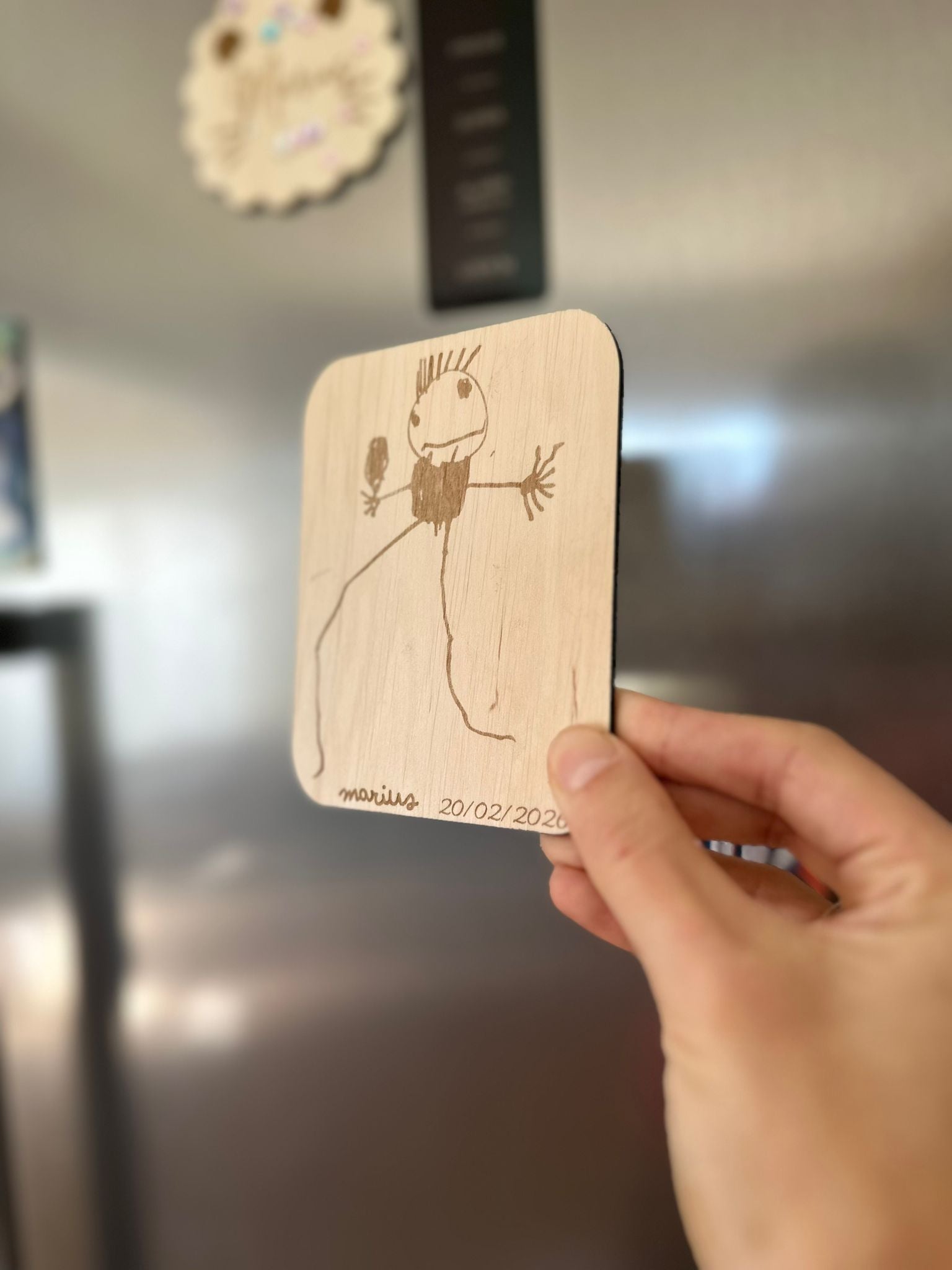 le dessin de votre enfant reproduit sur un magnet en bois pour un cadeau unique (tenu par une main d'adulte en face d'un réfrigérateur)
