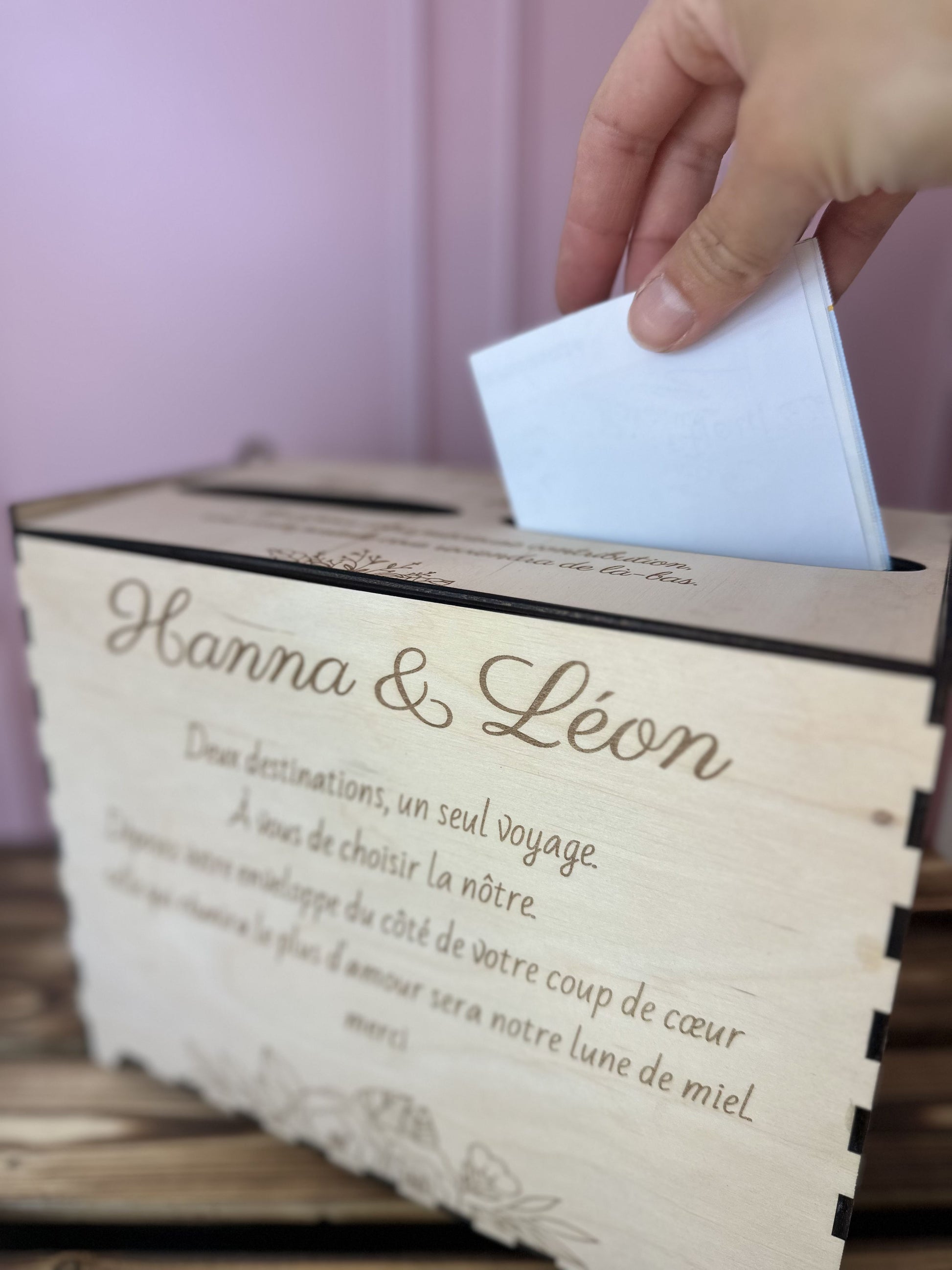 main glissant un chèque dans urne mariage en bois personnalisée "Hanna & Léon" et message gravé personnalisé.IL y a 2 entrées pour voter pour destination lune de miel. le fond est rose et la table sur laquelle est posée l'urne est en bois massif