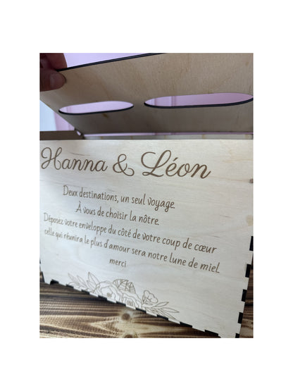 Urne mariage en bois personnalisée "Hanna & Léon" et message gravé personnalisé "deux destinations, un seul voyage. à vous de choisir la nôtre. Déposez votre enveloppe du côté de votre coup de coeur. celle qui réunira le plus d'amour sera notre lune de miel. merci" .IL y a 2 entrées pour voter pour destination lune de miel. le fond est rose et la table sur laquelle est posée l'urne est en bois massif le couvercle se soulève
