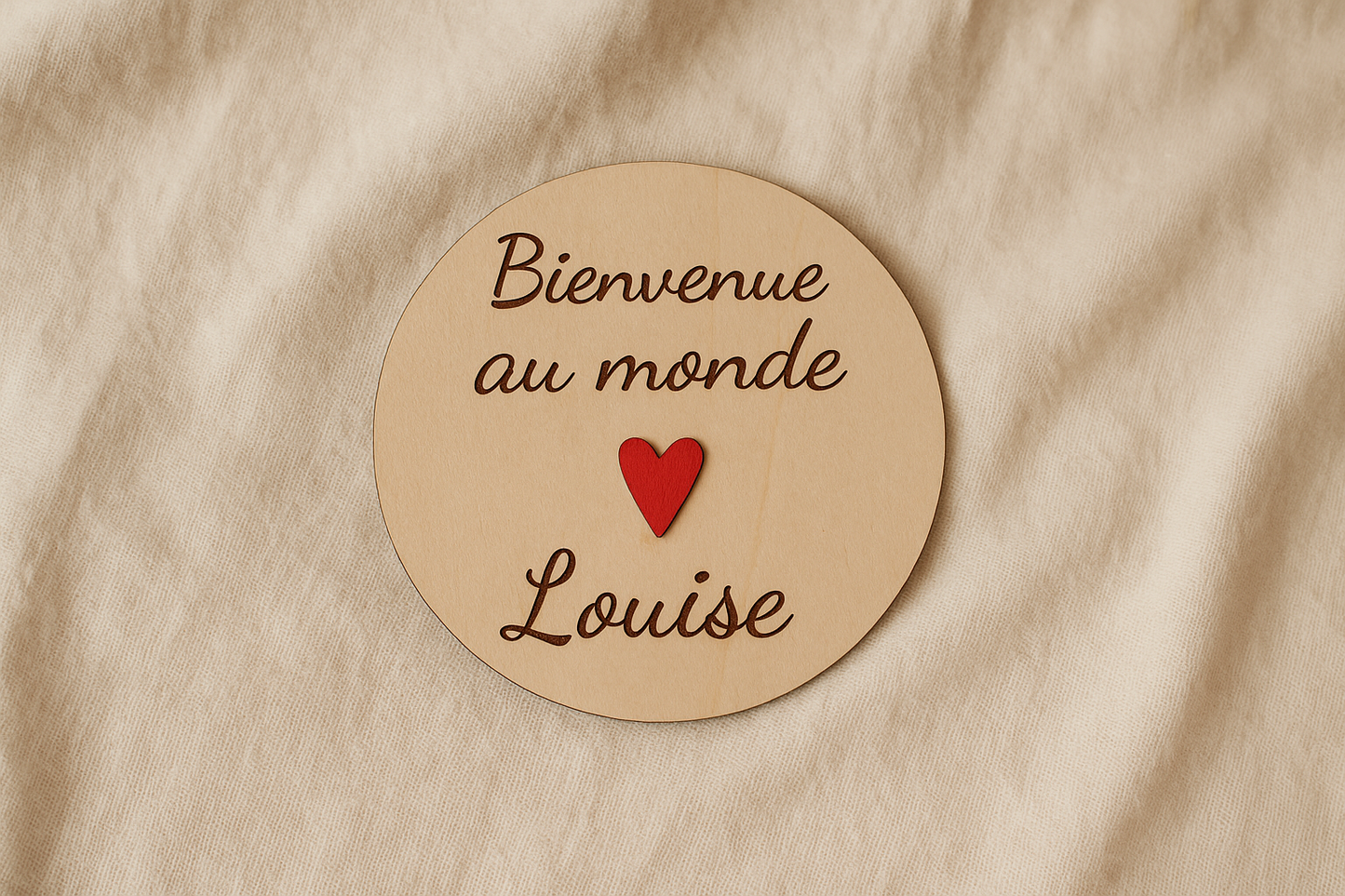 carte ronde en bois annonce naissance personnalisée avec le prénom de bébé Bienvenue au monde Louise" et avec un coeur rouge fixé au milieu. la carte en bois est posée sur un drap lin
