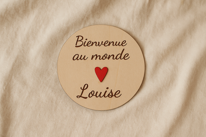 carte ronde en bois annonce naissance personnalisée avec le prénom de bébé Bienvenue au monde Louise" et avec un coeur rouge fixé au milieu. la carte en bois est posée sur un drap lin