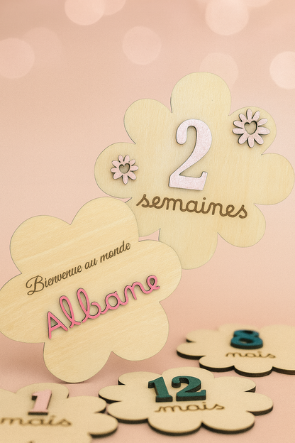 2 cartes en bois en forme de fleur : l'une gravée "Bienvenue au monde" avec le prénom "Albane" peint en rose et collé puis l'autre avec le numéro "2" peint en rose pâle et collé avec 2 fleurs de la même couleurs et "semaines" gravé. Posées en dessous : 3 cartes étapes, 2 vertes et 1 rose pâle; Le fond de la poto est rose pâle