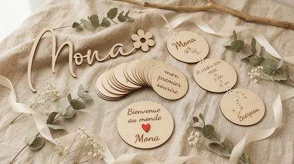 coffret de naissance personnalisé : prénom en bois avec fleur, lot de 34 cartes étapes, carte de bienvenue au monde personnalisée, suspension de naissance avec 3 disques avec les infos de naissance de bébé. Cadeaux de naissance en bois posés sur un linge en lin, avec des branches d'eucalyptus et des rubans