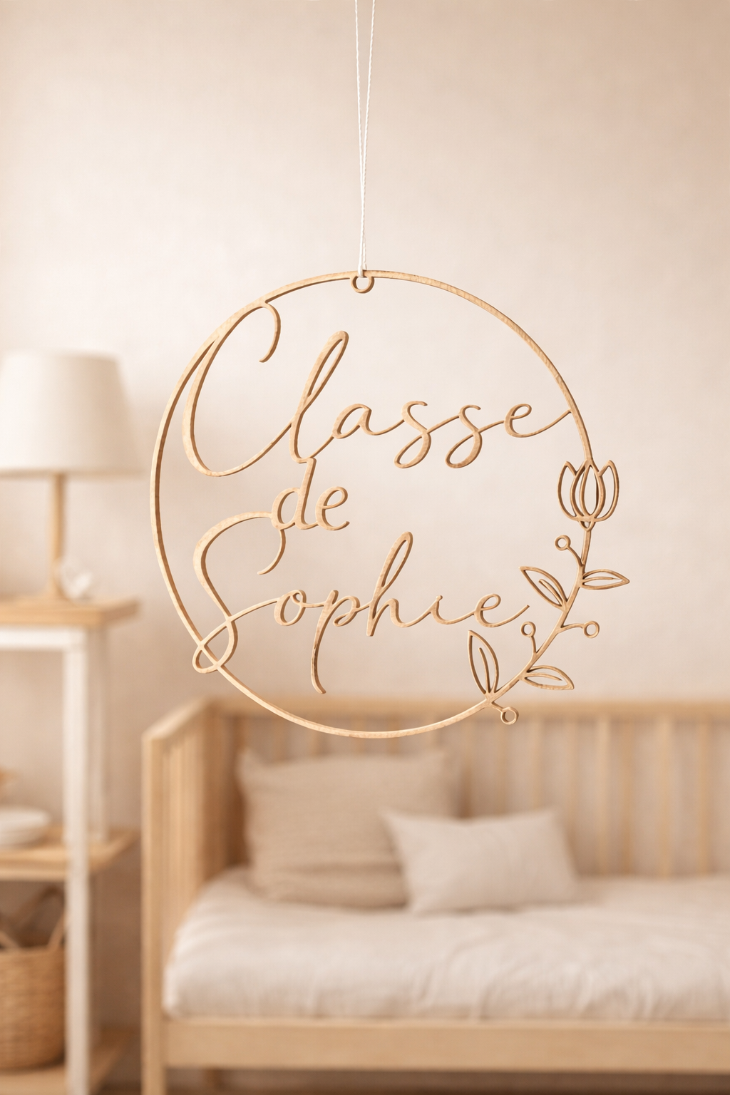 Couronne personnalisée en bois découpé au laser “Classe de Sophie”, suspendue par une ficelle, photographiée dans une chambre d’enfant minimaliste aux tons beige, avec lit en bois et lumière naturelle douce en arrière-plan.