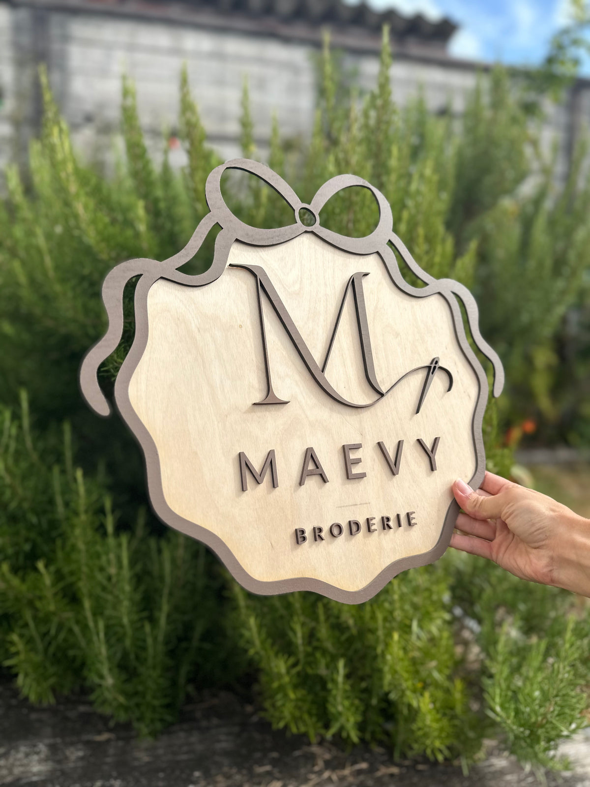 création en bois avec le logo de votre entreprise en 3 dimensions : MAEVY BRODERIE peint en marron et encadré d'un ruban en bois peint en marron. La pancarte logo en bois est tenue par une main devant du romarin