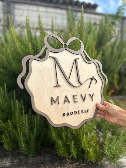 création en bois avec le logo de votre entreprise en 3 dimensions : MAEVY BRODERIE peint en marron et encadré d'un ruban en bois peint en marron. La pancarte logo en bois est tenue par une main devant du romarin