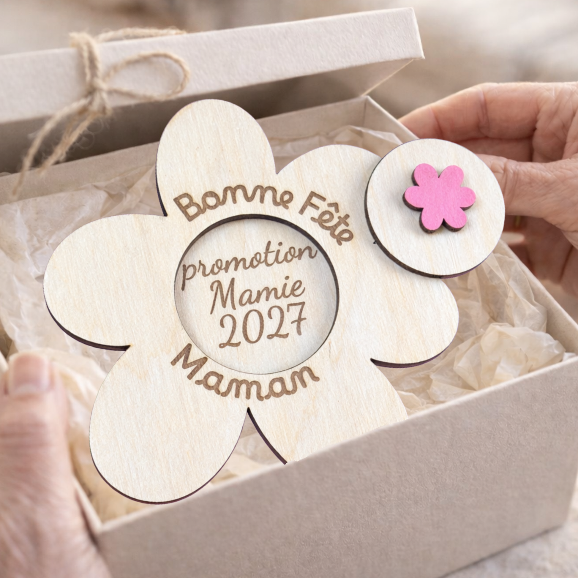 annonce grossesse en bois en forme de fleur. message gravé dessus "bonne fête maman" et le message à découvrir sous le bouton soulever grâce à une fleur rose en bois "promotion mamie 2027"; la fleur en bois est dans une boite en carton tapissée de papier de soi beige et tenue dans les mains un peu ridées d'une femme