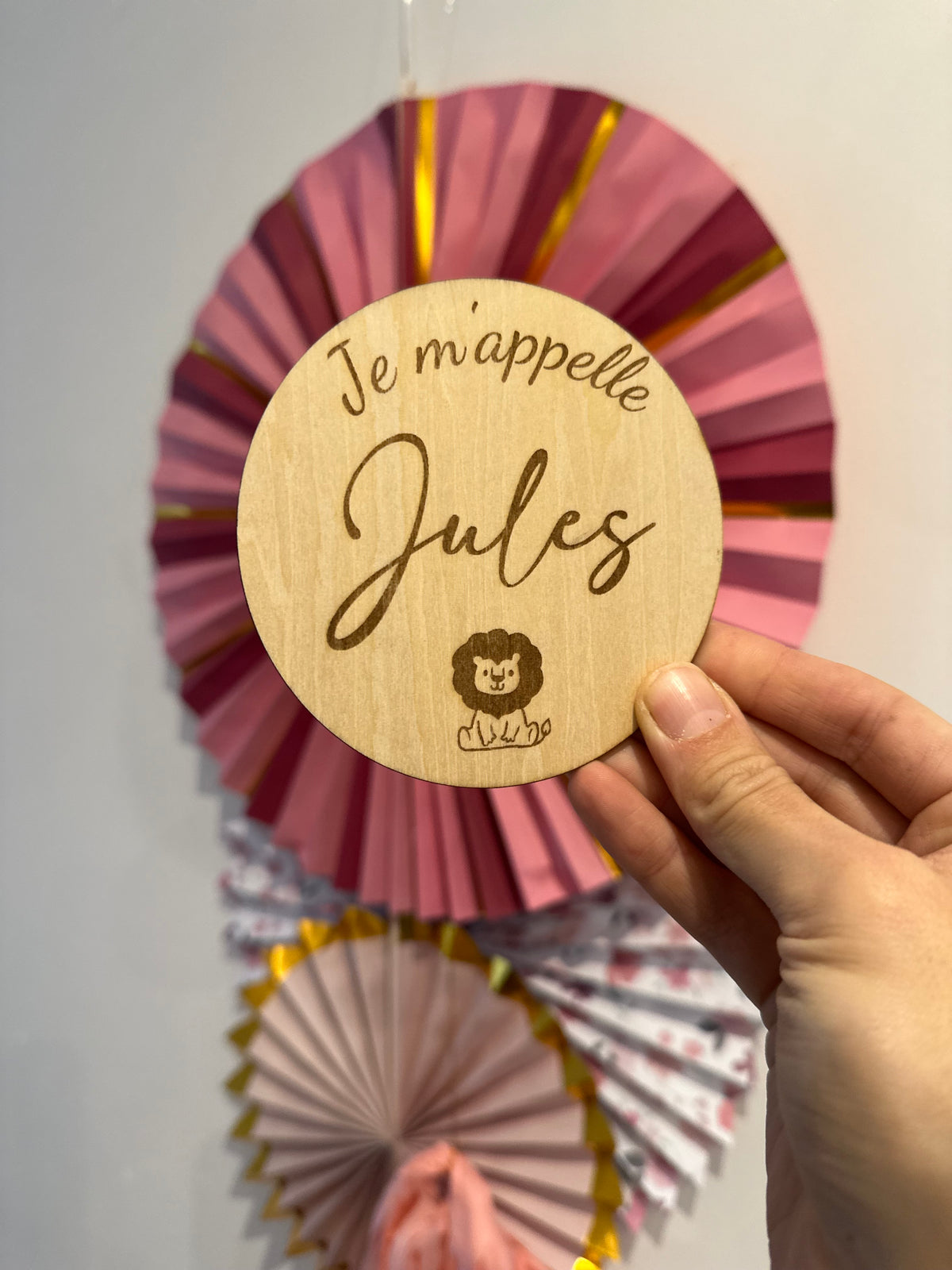 je m'appelle annonce naissance bébé personnalisée avec son prénom met un motif au choix, ici lion. La carte ronde en bois est tenue par une main en face d'une décoration rosace en papier rose