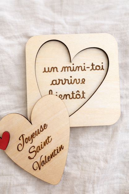 annonce grossesse spéciale saint Valentin en bois et en forme de coeur posé sur un tissu en lin clair : message gravé "un mini-toi arrive bientôt" et sur le couvercle à soulever "joyeuse saint Valentin"