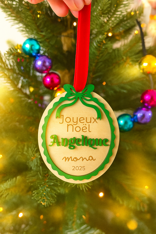 Suspension de Noël personnalisée en bois gravé, accrochée à un sapin décoré. L’ornement rond, orné d’un nœud vert et d’un ruban rouge, porte l’inscription “Joyeux Noël Angélique – Mona 2025”. En arrière-plan, les boules colorées et les guirlandes lumineuses créent une ambiance féerique et chaleureuse, typique de la collection artisanale Chou Rose