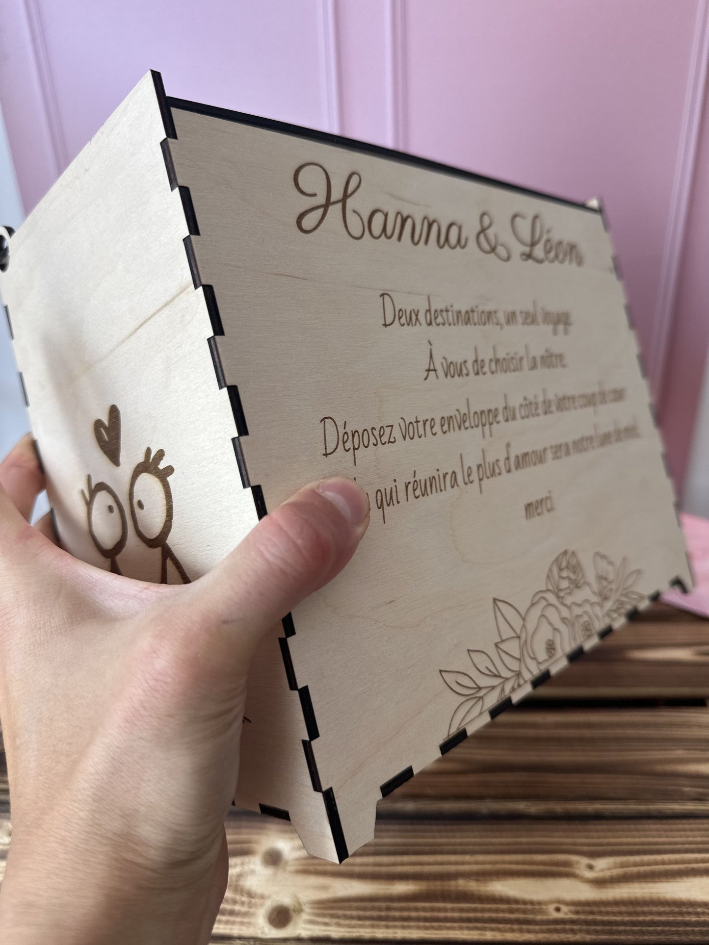 urne en bois personnalisée pour le mariage d'Hanna & Léon avec un message personnalisé gravé dessus "deux destinations, un seul voyage. a vous de choisir la nôtre. déposez votre enveloppe du côté de votre coup de coeur, celle qui réunira le plus d'amour sera notre lune de miel. merci", l'urne est tenue par une main, la table est en bois massif et le fond rose 
