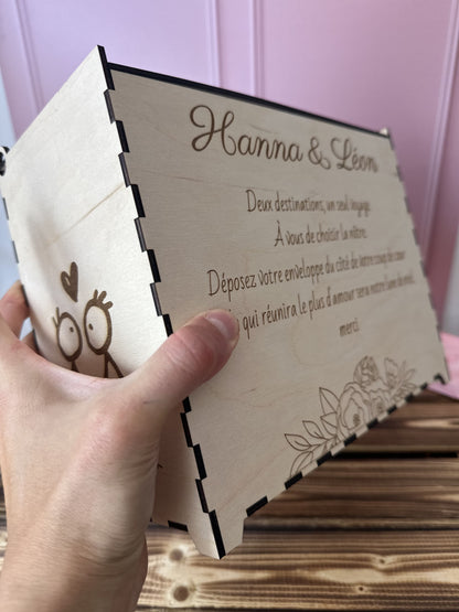 urne en bois personnalisée pour le mariage d'Hanna & Léon avec un message personnalisé gravé dessus "deux destinations, un seul voyage. a vous de choisir la nôtre. déposez votre enveloppe du côté de votre coup de coeur, celle qui réunira le plus d'amour sera notre lune de miel. merci", l'urne est tenue par une main, la table est en bois massif et le fond rose 