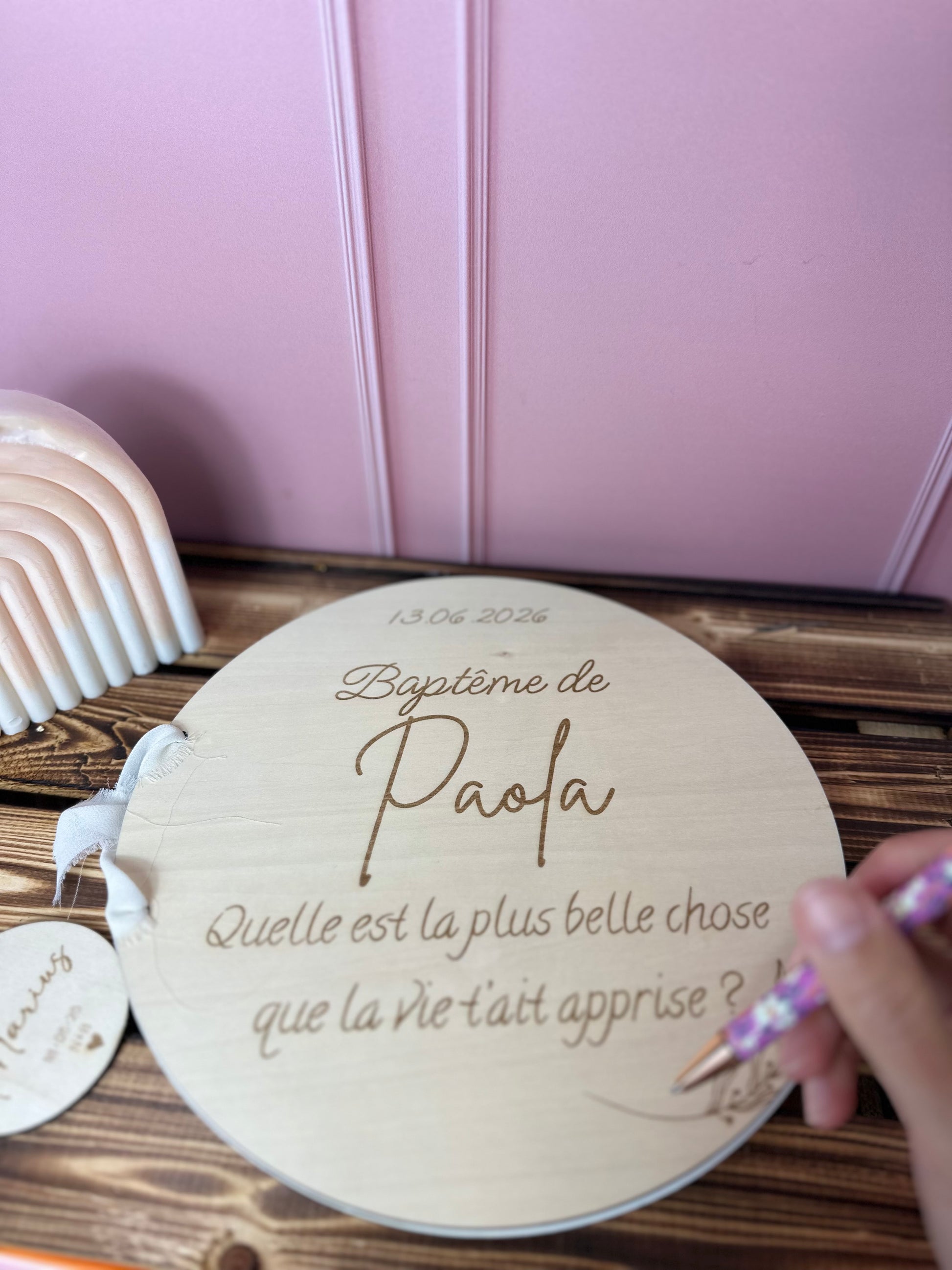 livre d'or baptême personnalisé avec un message ou une question gravé sur bois "baptême de Paola - quelle est la plus belle chose que la vie t'ait apprise ?" livre d'or décoré de motifs végétaux. posé sur une table en bois, une main tient un stylo rose dans les mains