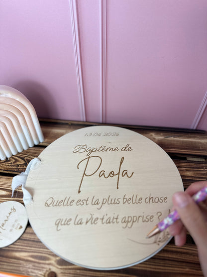 livre d'or baptême personnalisé avec un message ou une question gravé sur bois "baptême de Paola - quelle est la plus belle chose que la vie t'ait apprise ?" livre d'or décoré de motifs végétaux. posé sur une table en bois, une main tient un stylo rose dans les mains