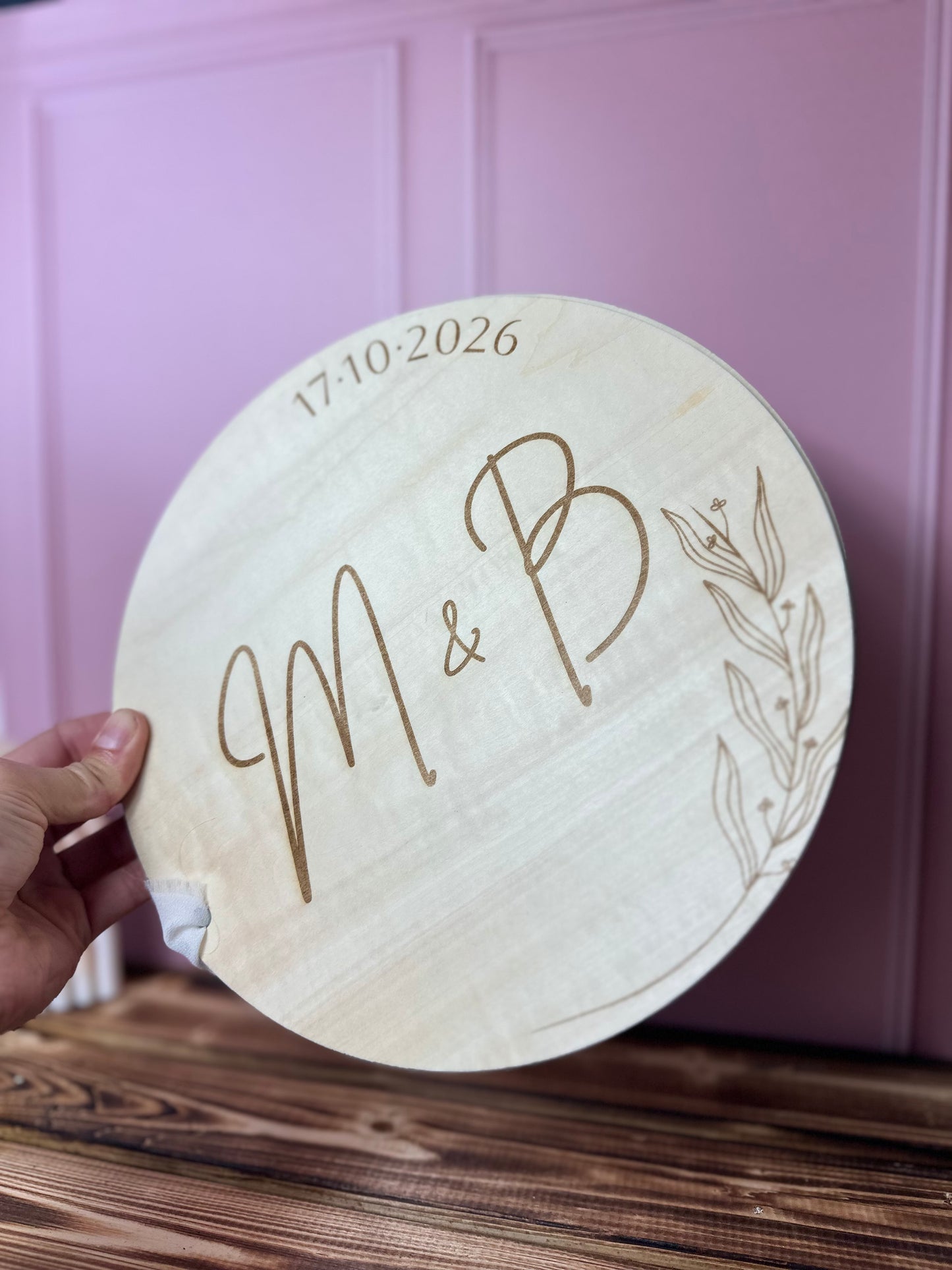 livre d'or en bois gravé avec les initiales pour mariage champêtre, et la date du mariage. le livre d'or personnalisé est rond est relié par un ruban blanc<; une main le tien au dessus d'une table en bois