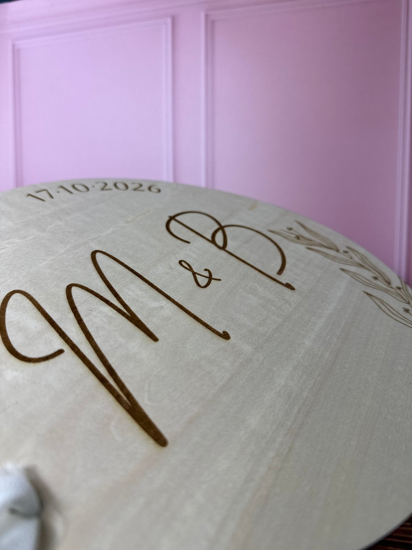 détails de la gravure personnalisée du livre d'or en bois gravé avec les initiales, les prénoms des mariés et la date du mariage. le livre d'or personnalisé est rond est relié par un ruban, fond mur rose