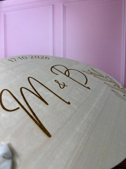 détails de la gravure personnalisée du livre d'or en bois gravé avec les initiales, les prénoms des mariés et la date du mariage. le livre d'or personnalisé est rond est relié par un ruban, fond mur rose