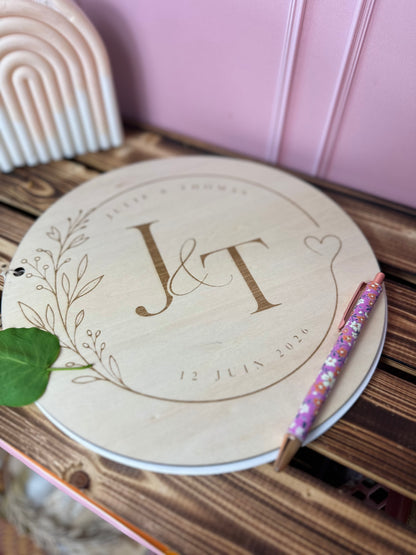 un livre d'or personnalisé en bois est posé sur une table en bois; les initiales des mariés (j&t) GRAVÉS en grand sur la couverture ainsi que la date et des éléments végétaux<; un stylo rose fleuri est posé sur le livre. en arrière plan une bougie en forme d'arche orangée et blanche