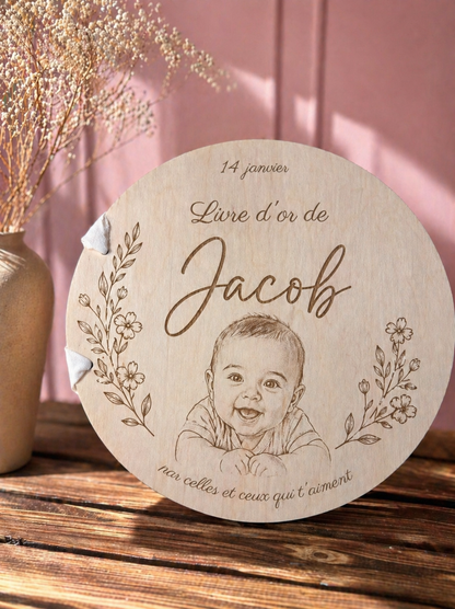 Livre d’or en bois personnalisé avec prénom et date gravés, portrait bébé gravé,  pages rondes, reliure avec un ruban blanc, posé sur une table en bois, fond rose éclairé par le soleil