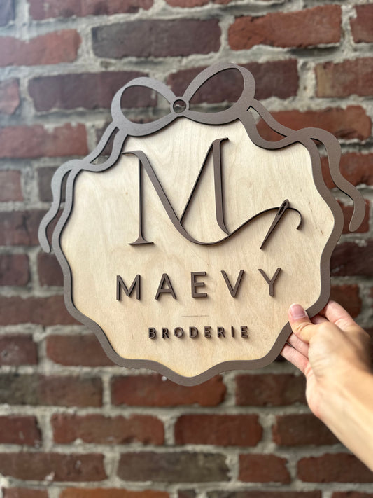 envoyez moi votre logo et nous en faisons une pancarte en bois en 3 dimensions avec la couleur de votre choix ! MAEVY BRODERIE en marron avec ruban marron tenue par une main devant un mur de briques rouges