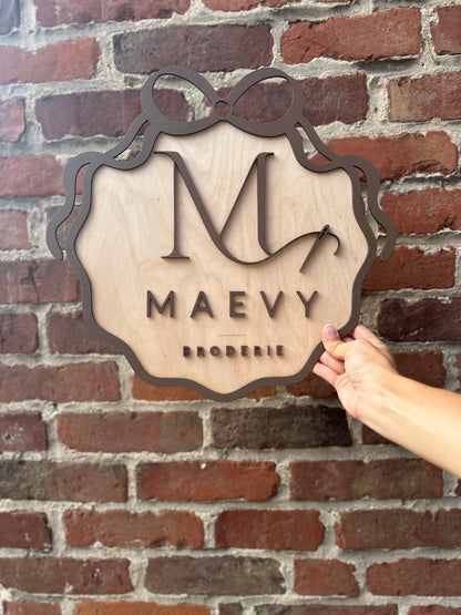 le logo de MAEY BRODERIE en 3 dimensions : logo découpé et peint puis collé sur une plaque en bois encadré d'un ruban en bois peint en marron. Le logo de la marque est tenue devant un mur de briques 