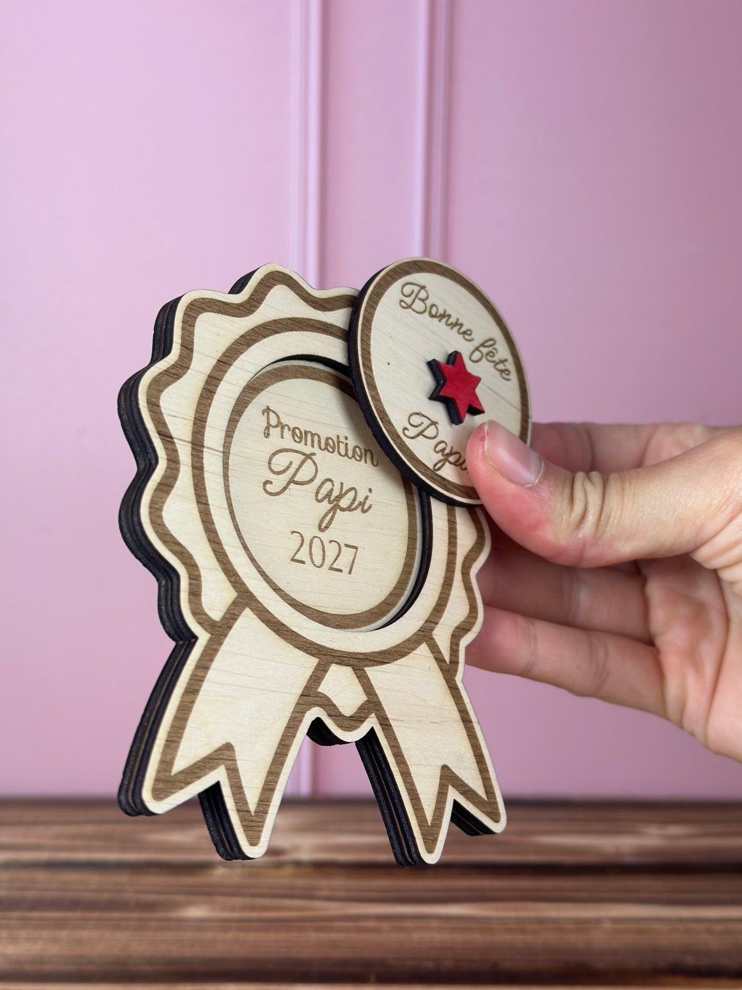 annonce grossesse  à découvrir pour Papi 2027 en forme de Médaille en bois gravée 
avec couvercle Bonne fête Papa à soulever - Chou Rose, tenue par une main féminine, fond : mur rose poudré, table : boid massif