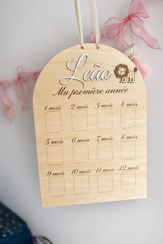 une pancarte personnalisée pour la première année de bébé avec son prénom peint en blanc et fixé "Leap" et un petit lion. il y a des emplacements pour les photos de sa première année. le panneau premier anniversaire est suspendu par un ruban crème, devant un mur décoré de rubans roses, 