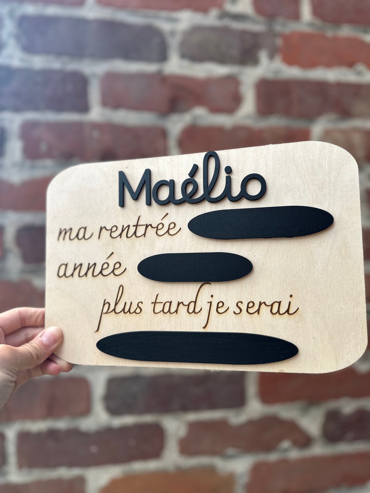 panneau en bois pour la rentrée à l'école personnalisé avec le prénom. 
Gravé : "ma rentrée :" - "année :" -"plus tard je serai : " et des ardoises pour remplir à la craie 
ici MAÉLIO en noir 

la pancarte est tenue par une main de femme, en fond des briques rouges 

