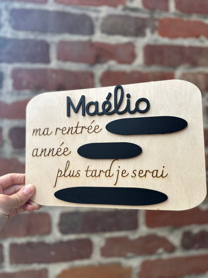 panneau en bois pour la rentrée à l'école personnalisé avec le prénom. 
Gravé : "ma rentrée :" - "année :" -"plus tard je serai : " et des ardoises pour remplir à la craie 
ici MAÉLIO en noir 

la pancarte est tenue par une main de femme, en fond des briques rouges 

